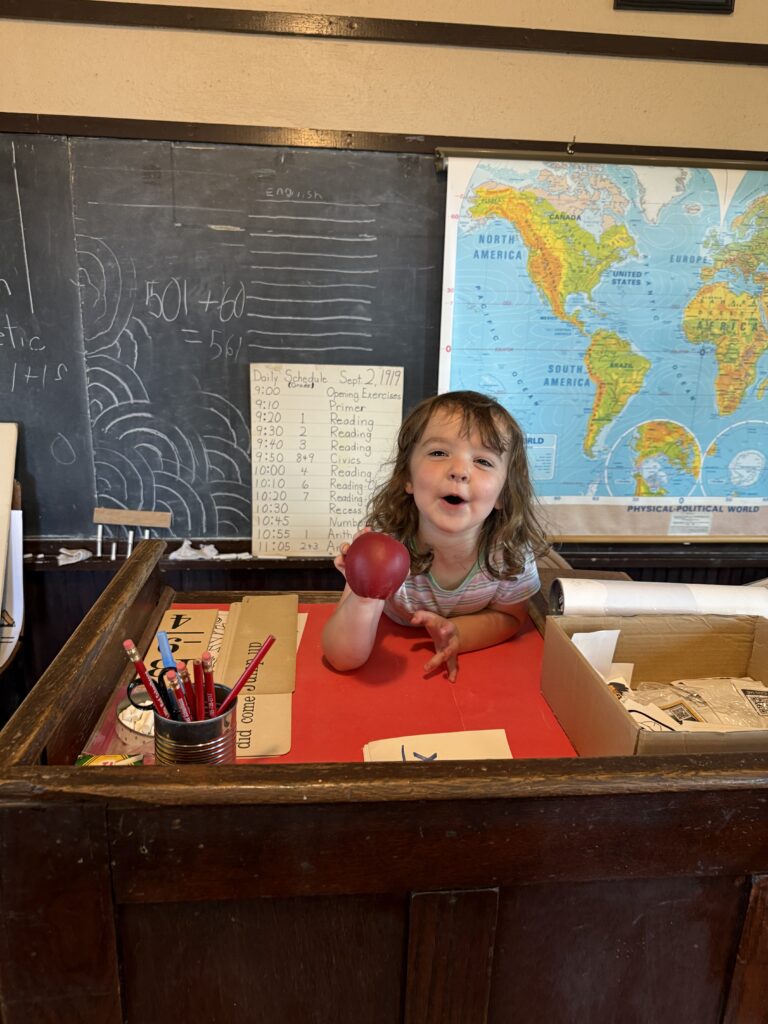 Girl holding apple at teacher's desk.